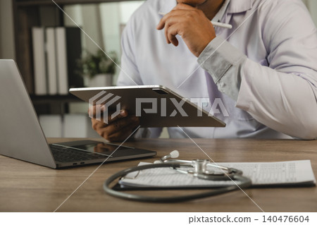 Healthcare professional reviewing patient records on a tablet beside a laptop and stethoscope at a medical desk, illustrating clinical documentation, digital healthcare workflow, patient information Healthcare professional reviewing patient records on a tablet beside a laptop and stethoscope at a medical desk, illustrating clinical documentation, digital healthcare workflow, patient information 140476604