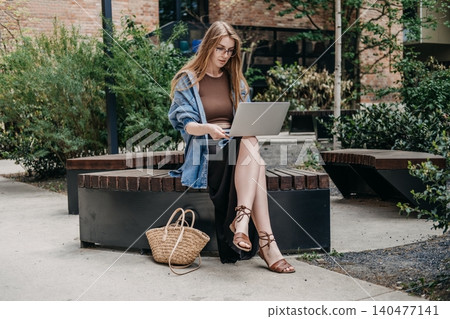 Woman operates laptop while sitting on public bench in outdoor courtyard. Freelance career outdoor, young professional working, flexible working hours. Woman operates laptop while sitting on public bench in outdoor courtyard. Freelance career outdoor, young professional working, flexible working hours. 140477141