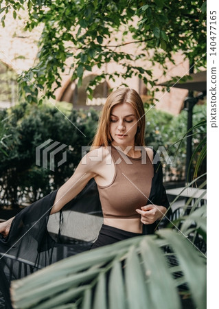 Woman adjusts lightweight fabric shirt while standing in green outdoor terrace. Clean look aesthetic, healthy lifestyle woman, serene facial expression. Woman adjusts lightweight fabric shirt while standing in green outdoor terrace. Clean look aesthetic, healthy lifestyle woman, serene facial expression. 140477165