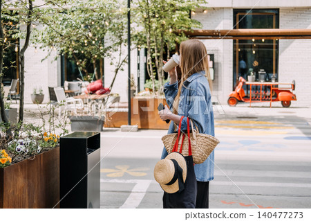 Woman standing outdoors sips beverage while holding smartphone and carrying woven tote bag. Sustainable living, urban lifestyle, eco friendly choices. 140477273