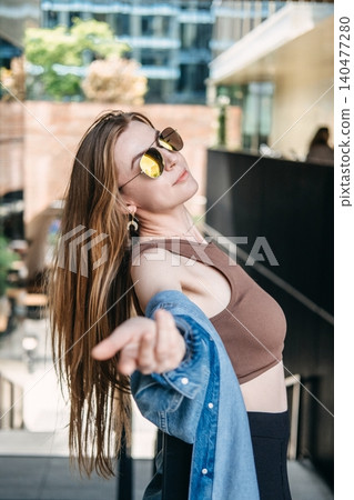 Woman extends hand forward while posing outside in city environment. Authentic city break, mindful moment outdoors, escaping busy street. Woman extends hand forward while posing outside in city environment. Authentic city break, mindful moment outdoors, escaping busy street. 140477280