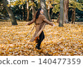 Woman walks through park filled with fallen autumn leaves while looking back. Low carbon footprint vacation, autumn outdoor recreation industry, sustainable tourism experiences. 140477359