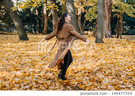 Woman walks through park filled with fallen autumn leaves while looking back. Low carbon footprint vacation, autumn outdoor recreation industry, sustainable tourism experiences. 140477359