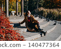 Woman sits on stone park steps surrounded by birds in sunlight. Mindful walking outdoor therapy, slow living autumn routine, connecting with nature footprints. 140477364