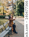 Woman sits on bench in public park while taking selfie or recording video with smartphone. User generated content UGC production, mobile photography in nature, social media marketing imagery. 140477366