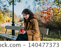 Woman sits on park bench holding insulated mug during autumn day. Mental health break, nature therapy, outdoor routine. 140477373
