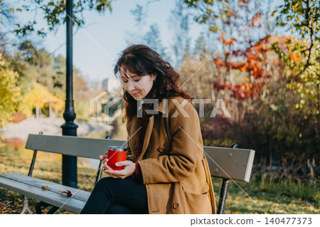 Woman sits on park bench holding insulated mug during autumn day. Mental health break, nature therapy, outdoor routine. Woman sits on park bench holding insulated mug during autumn day. Mental health break, nature therapy, outdoor routine. 140477373