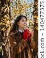 Woman stands in woods holding beverage container with both hands while looking upward. Mindfulness outdoor breathing exercise, positive thinking mental health, peaceful nature walk reflection. 140477375
