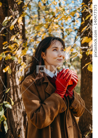 Woman stands in woods holding beverage container with both hands while looking upward. Mindfulness outdoor breathing exercise, positive thinking mental health, peaceful nature walk reflection. Woman stands in woods holding beverage container with both hands while looking upward. Mindfulness outdoor breathing exercise, positive thinking mental health, peaceful nature walk reflection. 140477375