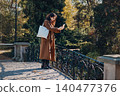 Woman stands on pedestrian bridge holding smartphone to take photograph. Taking photo with smartphone camera, outdoor content creator lifestyle, millennial blogger capturing moment. 140477376