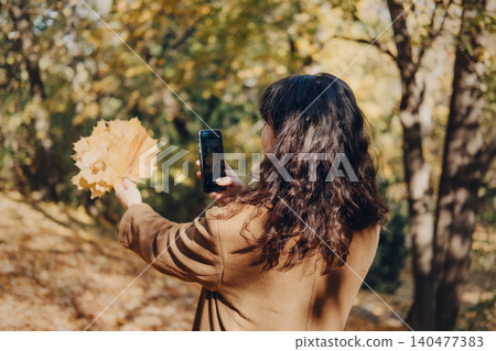 Person takes smartphone picture of yellow autumn leaves while smiling in park. Social media influencer lifestyle, autumn blogging content, lifestyle vlogger in park. Person takes smartphone picture of yellow autumn leaves while smiling in park. Social media influencer lifestyle, autumn blogging content, lifestyle vlogger in park. 140477383