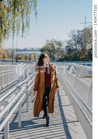 Woman walks on modern pedestrian bridge walkway while looking sideways. Walkable city infrastructure, modern urban pedestrian zone, navigating city architecture, accessible urban environment. 140477385