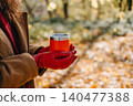 Person holds reusable beverage container in hands while standing in forest. Cozy hot drink outdoor comfort, winter transition seasonal marketing, warm beverage warming up hands. 140477388