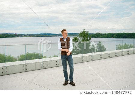 Middle-aged man stands on terrace overlooking lake rolling up sleeve of his shirt Middle-aged man stands on terrace overlooking lake rolling up sleeve of his shirt 140477772