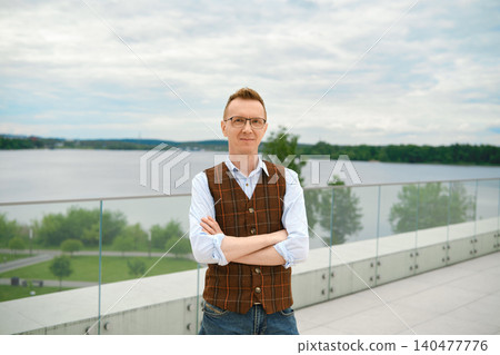 Middle-aged man stands on balcony of observation deck above lake in park with his arms crossed over his chest Middle-aged man stands on balcony of observation deck above lake in park with his arms crossed over his chest 140477776