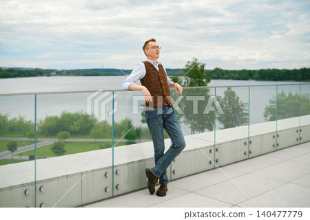 Middle-aged man in vest and jeans leans against glass railing with river in background Middle-aged man in vest and jeans leans against glass railing with river in background 140477779