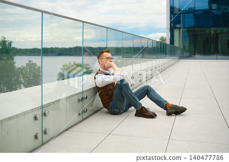 Thoughtful middle-aged man sitting on floor near glass railing beside river on cloudy day Thoughtful middle-aged man sitting on floor near glass railing beside river on cloudy day 140477786