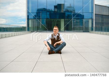 Middle-aged man sitting on ground outside modern building with glass walls during cloudy day Middle-aged man sitting on ground outside modern building with glass walls during cloudy day 140477788
