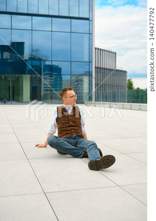 Middle-aged man in vest and jeans sits on ground in urban area near modern buildings during daytime 140477792