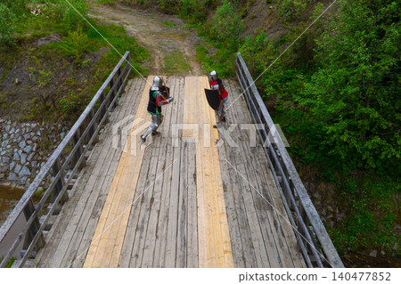 People actors dressed as medieval knights spar with swords and shields on a weathered bridge in a natural, wooded setting 140477852