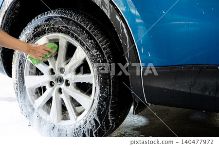 A person is washing a car tire with a green brush. Tire is covered in water and soap. Blue car wash with white soap foam. Auto care service. Car cleaning service concept. Vehicle cleaning service. A person is washing a car tire with a green brush. Tire is covered in water and soap. Blue car wash with white soap foam. Auto care service. Car cleaning service concept. Vehicle cleaning service. 140477972