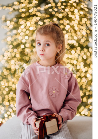 Girl holding a gift sits in front of a Christmas tree with lights Girl holding a gift sits in front of a Christmas tree with lights 140478021