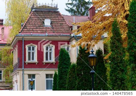 A historic European building with red brick accents and ornate windows is framed by vibrant yellow autumn leaves and lush green cypress trees A historic European building with red brick accents and ornate windows is framed by vibrant yellow autumn leaves and lush green cypress trees 140478565