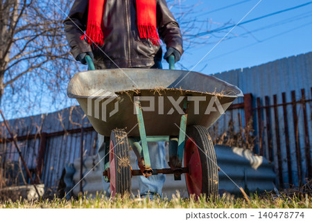 Man gardener pushing wheelbarrow in yard during winter. Male worker using equipment for yard cleaning. Concept of home maintenance, garden preparation, hard labor and seasonal outdoor work. Man gardener pushing wheelbarrow in yard during winter. Male worker using equipment for yard cleaning. Concept of home maintenance, garden preparation, hard labor and seasonal outdoor work. 140478774