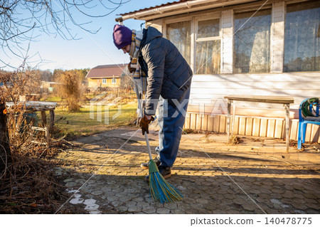 man cleaning backyard patio with broom. Person sweeping stone tile path near countryside house. Yard maintenance and autumn garden preparation. Outdoor chores in residential area. man cleaning backyard patio with broom. Person sweeping stone tile path near countryside house. Yard maintenance and autumn garden preparation. Outdoor chores in residential area. 140478775