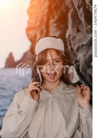 Woman coastal sunset playful young woman wearing a ushanka hat sticking her tongue out near rocky cliffs by the sea Woman coastal sunset playful young woman wearing a ushanka hat sticking her tongue out near rocky cliffs by the sea 140479674