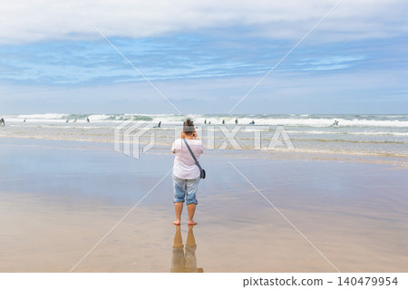 A woman stands on a wet, reflective sandy beach, taking a photo of surfers riding ocean waves. This bright scene evokes freedom, travel wanderlust, and peaceful coastal vacation vibes. A woman stands on a wet, reflective sandy beach, taking a photo of surfers riding ocean waves. This bright scene evokes freedom, travel wanderlust, and peaceful coastal vacation vibes. 140479954