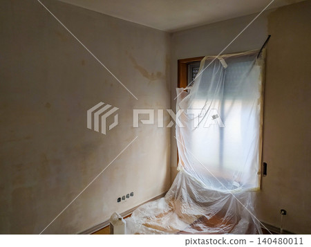 Interior room undergoing renovation with stained plaster walls and a window covered in protective plastic sheeting, capturing the messy preparation phase of home improvement projects. Interior room undergoing renovation with stained plaster walls and a window covered in protective plastic sheeting, capturing the messy preparation phase of home improvement projects. 140480011