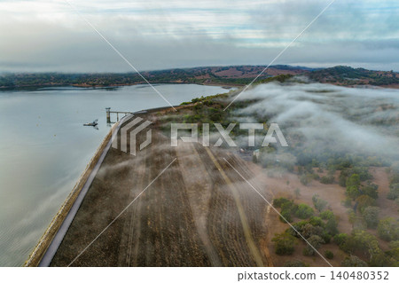 Aerial image of the Glenbawn Dam wall with morning fog drifting through the surrounding valley in NSW Australia. Drone photograph captures the reservoir, rugged mountain landscape Aerial image of the Glenbawn Dam wall with morning fog drifting through the surrounding valley in NSW Australia. Drone photograph captures the reservoir, rugged mountain landscape 140480352