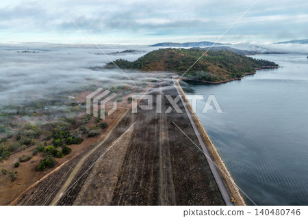 Vertical Aerial view of the Glenbawn Dam wall leading towards the surrounding mountain landscape with fog rolling through the valley in NSW Australia. Drone photograph captures atmospheric low cloud Vertical Aerial view of the Glenbawn Dam wall leading towards the surrounding mountain landscape with fog rolling through the valley in NSW Australia. Drone photograph captures atmospheric low cloud 140480746