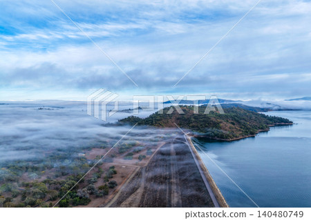 Vertical Aerial view of the Glenbawn Dam wall leading towards the surrounding mountain landscape with fog rolling through the valley in NSW Australia. Drone photograph captures atmospheric low cloud Vertical Aerial view of the Glenbawn Dam wall leading towards the surrounding mountain landscape with fog rolling through the valley in NSW Australia. Drone photograph captures atmospheric low cloud 140480749