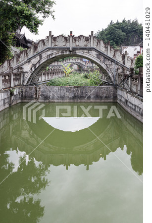 an ornate, weathered stone arch bridge adorned with intricate bas-relief carvings spans gracefully over a canal, casting a flawless, semi-circular reflection onto the calm, greenish waters below. an ornate, weathered stone arch bridge adorned with intricate bas-relief carvings spans gracefully over a canal, casting a flawless, semi-circular reflection onto the calm, greenish waters below. 140481029