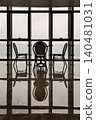 Three elegant woven chairs rest in quiet silhouette against a grand grid window, their elegant forms captured in a flawless, mirror-like reflection upon the dark, polished floor below. 140481031