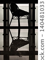 a single, high-backed bistro chair is captured in a striking silhouette against a structural window frame, its geometric lines casting a flawless, liquid-like reflection onto the dark, polished 140481033