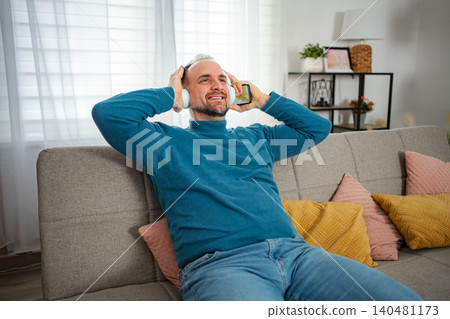 Man relaxing on sofa listening to music with headphones Man relaxing on sofa listening to music with headphones 140481173