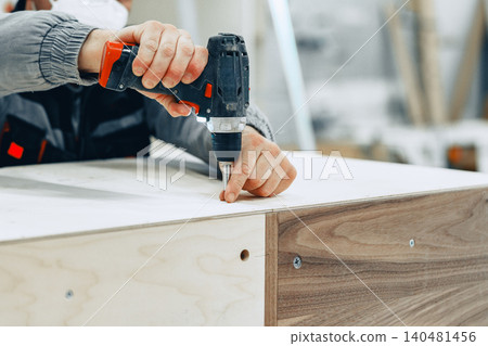 Worker uses drill in a furniture workshop to assemble wood pieces on a project 140481456