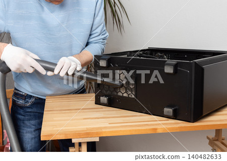 A 53-year-old man uses a vacuum to clean dust from a computer system unit on a wooden table in a home office. The setting is tidy and organized A 53-year-old man uses a vacuum to clean dust from a computer system unit on a wooden table in a home office. The setting is tidy and organized 140482633