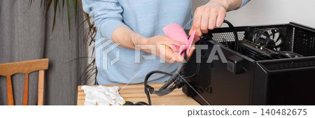 A 53-year-old man uses a cloth to clean dust from a computer system unit. He works at a wooden table in a room with plants nearby. Tools are set out for the task A 53-year-old man uses a cloth to clean dust from a computer system unit. He works at a wooden table in a room with plants nearby. Tools are set out for the task 140482675