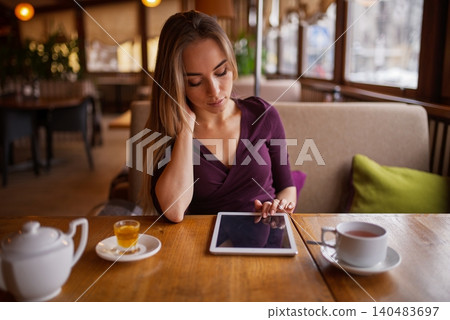 A beautiful blonde woman using a digital tablet at a wooden table with tea inside a cozy cafe A beautiful blonde woman using a digital tablet at a wooden table with tea inside a cozy cafe 140483697