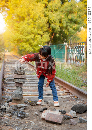 Curious little girl looks at a stack of balanced stones next to an old railway track in nature. Curious little girl looks at a stack of balanced stones next to an old railway track in nature. 140483734