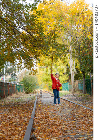 A happy young girl plays with a long wooden stick while standing under autumn trees near a railway track. A happy young girl plays with a long wooden stick while standing under autumn trees near a railway track. 140483737