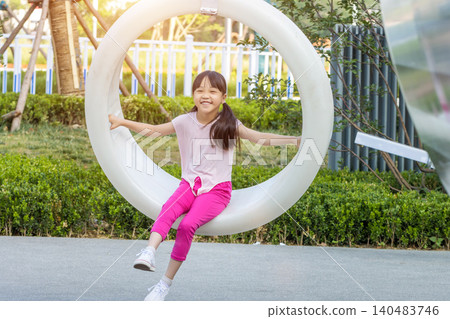 Happy elementary girl sitting in a white suspended plastic circle swing at an outdoor park, looking at the camera with a joyful smile Happy elementary girl sitting in a white suspended plastic circle swing at an outdoor park, looking at the camera with a joyful smile 140483746