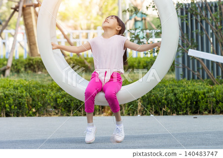 A cheerful young girl sitting on a large white circular swing at an outdoor playground. A cheerful young girl sitting on a large white circular swing at an outdoor playground. 140483747