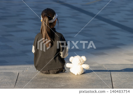 Sad and lonely little girl crying and hugging teddy bear standing by the wall outdoors Sad and lonely little girl crying and hugging teddy bear standing by the wall outdoors 140483754