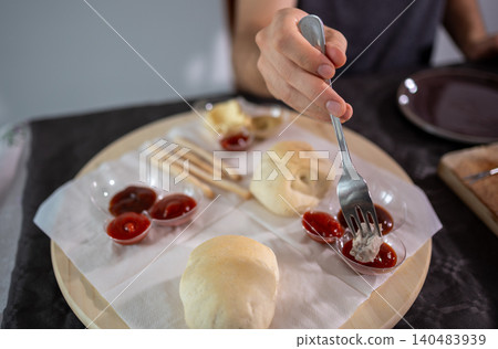 Person using a fork to dip a piece of cooked meat into a small serving of red sauce, with other sauces, bread, and breadsticks arranged on a wooden platter Person using a fork to dip a piece of cooked meat into a small serving of red sauce, with other sauces, bread, and breadsticks arranged on a wooden platter 140483939