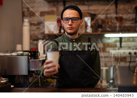 A male barista standing behind a bar counter holding a paper coffee cup 140483943